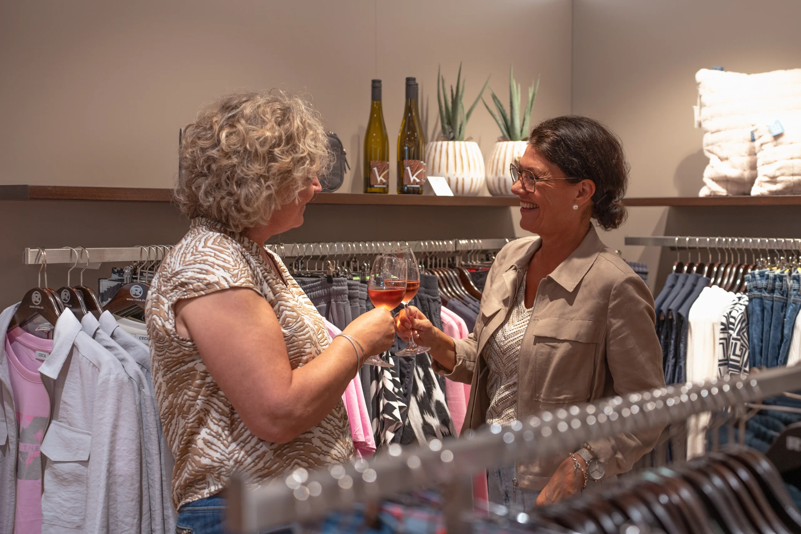 Zwei Frauen stoßen mit Wein an im Modehaus Rieke zwischen moderner Damenmode